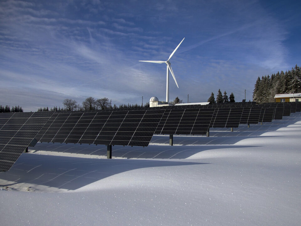 Ein Feld mit Solarzellen, im Hintergrund ein Windrand im schneereichen Winter