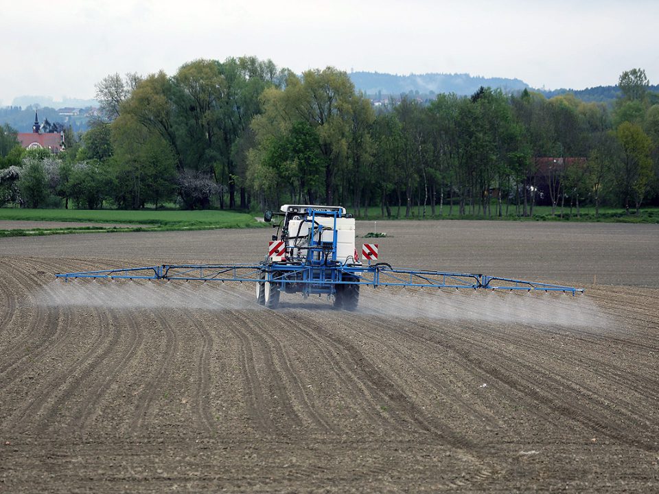 Traktor fährt düngend über einen Acker. Im Hintergrund Laubbäume, Gebäude und Berge